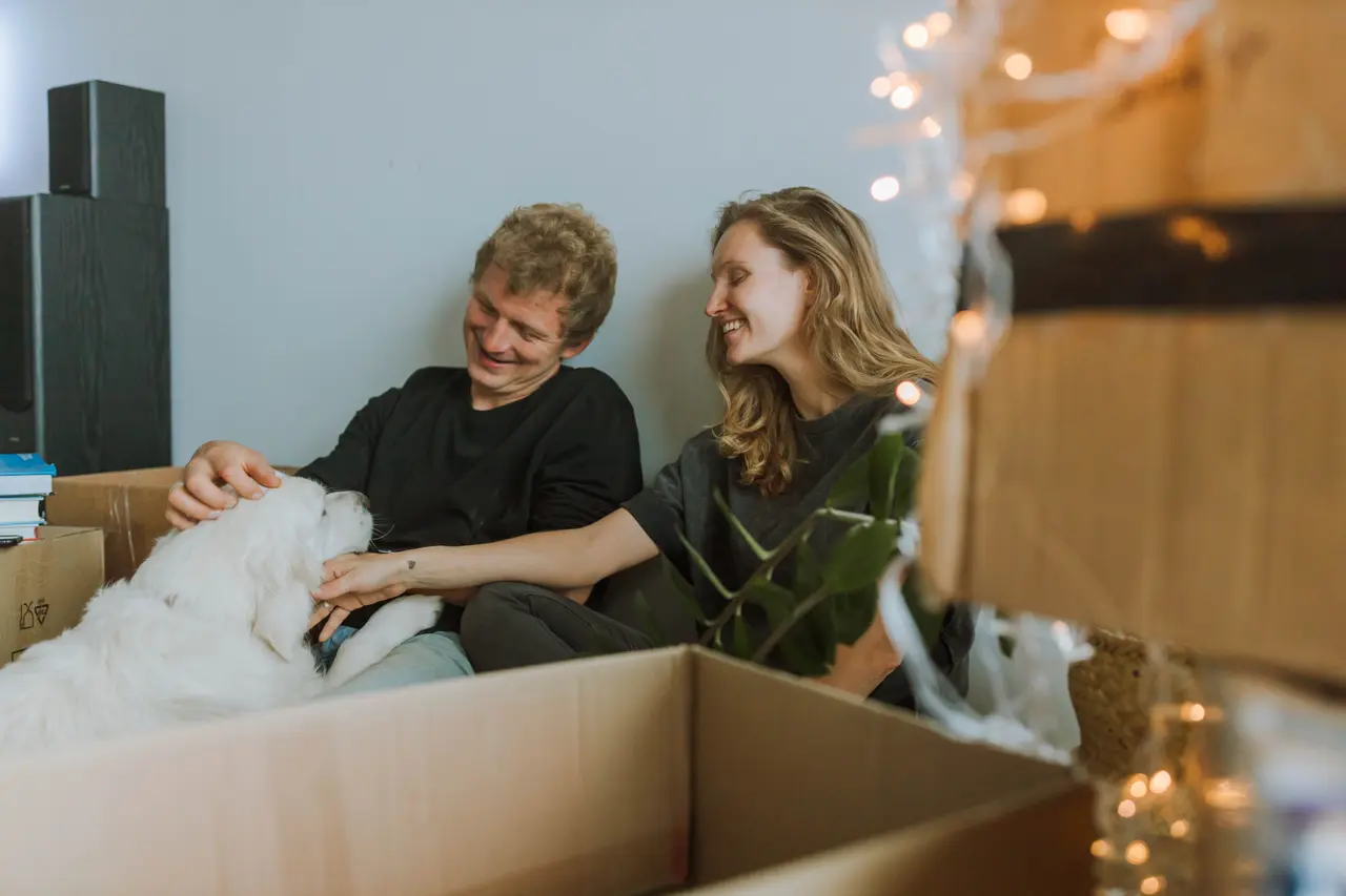 A smiling couple sits among open moving boxes, petting their white dog